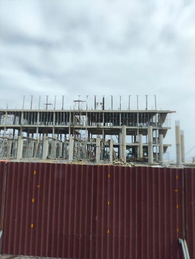 The image displays a multi-story reinforced concrete building under construction, located in Tuy Phong 4, Vietnam, viewed from behind a reddish-brown corrugated metal fence. The fence, featuring vertical corrugations and sporadic small yellow rectangular markings, occupies the lower third of the frame. Above the fence, the concrete structural skeleton of the building is prominently visible, consisting of multiple levels of columns and floor slabs. Exposed rebar extends vertically from the uppermost completed slab, indicating ongoing construction for additional stories. Metal scaffolding and support structures are integrated throughout the partially built edifice. No individuals, active machinery, or discernable construction activity are visible. The background is dominated by a uniformly overcast, grey sky.
