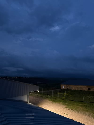 The image depicts a rural or semi-rural scene at dusk or during a stormy evening, likely in the vicinity of Sisian, Armenia. The sky is filled with dark, heavy clouds, contributing to a somber and dramatic atmosphere. In the foreground, the corrugated metal roof of a modern building is visible, with a bright, artificial light illuminating the walkway and the side of the structure. To the right, a more traditional, older stone building with a dark roof is situated further back. This building has at least one window lit from within, suggesting occupancy. A dirt path runs between the two buildings, bordered by green grass and a low wire fence. In the distance, a hilly landscape is discernible under the dim, overcast sky, with faint lights of what could be a settlement or scattered houses. The overall impression is one of quiet solitude and impending weather, set against a backdrop of rural Armenian topography.