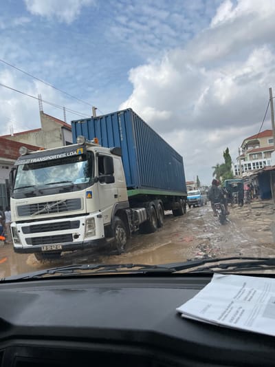 The image captures a street scene in Luanda, Angola, from the perspective of a vehicle's dashboard. The main subject is a large white Volvo truck with a blue shipping container loaded on its trailer, positioned on a muddy, unpaved road. The truck's cab has "VOLVO" prominently displayed on its grille, and its license plate "LD-51-42-EA" is visible. Text on the sun visor reads "RUA TRANSPORTES LDA". Part of the container's identification "ARTU 700" is also visible.
The road is extensively covered in large puddles and mud, suggesting recent rain. In the background, other vehicles, including a motorcycle with a rider, navigate the challenging terrain. Several people are visible in the distance and near the truck's front wheel. Buildings, some appearing residential or commercial, line the sides of the street, with a cloudy sky overhead. Overhead wires are also noticeable. The time of day is daytime, with bright but overcast conditions.
Inside the viewing vehicle, a white document with printed text is taped to the dashboard or windshield. Visible words on the document include "CONTRACTO" and a number "500.000". The scene depicts a bustling, everyday urban environment characterized by difficult road conditions.