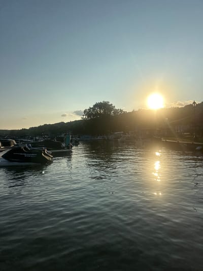 This is a photograph of a marina on a lake in Algonquin, Illinois. The sun is setting over the lake, casting a warm glow on the water and the surrounding trees. Several boats are docked in the marina, including a jet ski in the foreground. The water is calm, with gentle ripples reflecting the sunlight. The sky is a clear blue, with a few wispy clouds. The overall mood of the photograph is peaceful and serene.