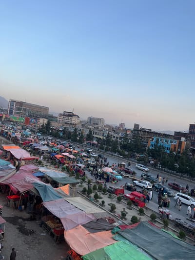 This is an aerial photograph of a bustling street market in Kabul, Afghanistan, likely in the late afternoon or early evening, as indicated by the warm light and clear sky. The scene is a vibrant, chaotic mix of commerce and daily life.

In the foreground, numerous market stalls are set up, covered by colorful tarps in shades of orange, pink, green, and blue. These stalls display a variety of goods, with piles of fruits, vegetables, and other produce visible on carts and tables. People are milling about the stalls, some appearing to be vendors and others customers. The ground beneath the stalls is uneven and appears to be a mix of dirt and paved areas.

A busy street runs parallel to the market, filled with a diverse array of vehicles including cars, vans, and possibly motorcycles. The traffic appears to be moderate, with vehicles moving in both directions. Pedestrians are also present on the sidewalks and near the street, some carrying bags or interacting with vendors.

In the background, the cityscape of Kabul rises. Several multi-story buildings are visible, ranging in architectural style from modern to older structures. Some buildings appear to be commercial or residential, with shops and offices on the ground floors. Trees are planted along the street and between some buildings, adding a touch of greenery to the urban environment. In the far distance, mountains frame the city, suggesting the mountainous terrain surrounding Kabul.

The overall impression is one of a lively, active urban center where everyday commerce and social interaction are taking place amidst the backdrop of the city. The image captures a sense of dynamism and the everyday rhythm of life in this part of Kabul.