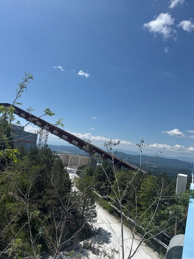 The image shows an elevated conveyor system, presumably for mining materials, located in Panagyurski kolonii, Bulgaria. The structure is red-brown with white-framed windows, supported by steel framework. In the background, a mountain range is visible under a blue sky with scattered clouds. In the foreground, there is green vegetation and a road. Part of a building structure is visible in the top right corner.