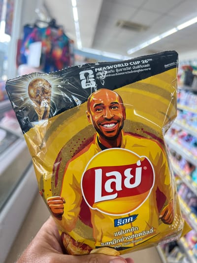 This media file features a close-up shot of a person's hand holding a bag of Lay's potato chips. The bag is predominantly yellow and features a stylized illustration of a smiling man, possibly a celebrity endorsement, with a red and white Lay's logo. The design also includes imagery related to the FIFA World Cup, with the FIFA logo and text mentioning "FIFA WORLD CUP 26TM". Some text is in Thai and English, with "Hot Chili Squid" clearly visible, indicating the flavor of the chips. The background appears to be an aisle in a store, likely a convenience store or supermarket, with shelves of products blurred in the distance. The lighting suggests an indoor, well-lit environment, typical of a retail setting. The focus is on the chip bag, highlighting its design and promotional elements.
