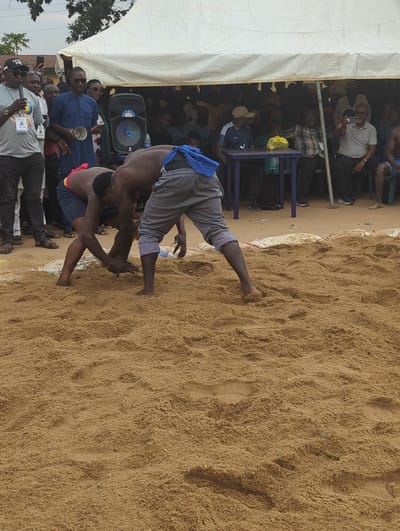 This image, taken in what is believed to be Kafanchan A, Nigeria, depicts a traditional wrestling match occurring in a sandy arena. Two shirtless men are engaged in a wrestling grapple at the center. Surrounding the arena, a crowd of spectators observes the match, some seated on chairs under a white tent structure. In the foreground, several individuals stand watching, including at least one person holding documents or promotional materials. A loudspeaker is situated near the tent. The background consists of additional spectators, with elements of the local environment visible in the distance.