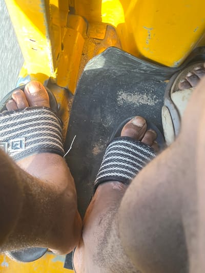 The image shows a close-up view of two feet wearing sandals, positioned inside a yellow vehicle that appears to be a tuk-tuk. The feet are resting on the floor of the vehicle, which is partly covered by a black mat. The sandals are black with a white striped pattern and a metallic emblem on the strap. Sand is visible on the black mat, suggesting an outdoor environment, possibly a dusty or sandy road.

The scene is shot from a high angle, looking down at the feet. The lighting suggests it is daytime, with sunlight illuminating the yellow exterior of the vehicle. The overall impression is that the person is a passenger or driver in a tuk-tuk in Kporva, Nigeria. There are no visible emotions conveyed by the image, as the focus is solely on the feet and the interior of the vehicle. No text is discernible in the image.