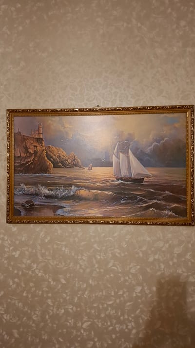 The image shows a framed painting of a seascape. A sailing ship with white sails is depicted in the middle ground of the painting, moving through choppy water. In the background, on the left, a castle or fortress sits atop a rocky cliff. The sky is filled with dramatic clouds, and the sun appears to be setting, casting a warm glow on the water and the ship. There are also smaller waves crashing on a rocky shore in the foreground. The painting is ornate with a golden frame. The painting is hung on a textured beige wall with a floral pattern. There is no indication of the city of Chemburka, Russia within the image itself.