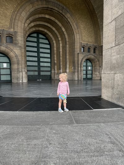 A young child with blonde hair, wearing a pink long-sleeved shirt, blue shorts, and sandals, stands in front of a large, ornate stone building with arched entrances and dark green doors. The child is holding a colorful toy in their right hand and a small plush bear is attached to their shorts. The ground in front of the building is paved with large stone slabs. The building is likely part of the National Basilica of the Sacred Heart in Koekelberg, Belgium, known for its distinctive Art Deco architecture. The scene appears to be an outdoor public space, possibly a plaza or esplanade, with the imposing facade of the basilica dominating the background. The lighting suggests it is daytime.