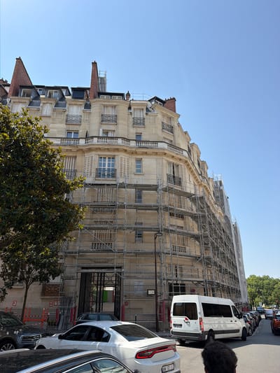 The image shows an apartment building undergoing renovation, with extensive scaffolding covering its facade. The building is light-colored stone with ornate architectural details, including balconies and decorative window grates. Multiple stories are visible, featuring numerous windows, some with blinds drawn. A large tree with dark green leaves is in the foreground, partially obscuring the lower part of the building.

In the street in front of the building, a white van is parked, its rear doors open. Several other vehicles are parked along the street, forming a line. The street appears to be paved, and there are trees and foliage visible in the background, suggesting an urban or suburban setting. The sky is clear and blue, indicating fair weather. The lighting suggests it is daytime, likely late morning or afternoon.

There is some visible text on the white van, including a license plate and possibly some small lettering. There are also signs on the scaffolding and on the side of the building, but they are too small to read clearly. The overall impression is of a busy urban scene where construction and daily life are coexisting.