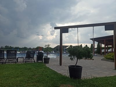 This image depicts an outdoor scene at a recreational facility, likely a resort or water park, in Sopot Urban Municipality, Serbia. The foreground features a neatly mowed lawn adjacent to a paved area. Several black lounge chairs are arranged near a swimming pool.
The swimming pool itself is visible, with clear blue water and some pool toys or inflatables present. In the mid-ground, a wooden structure with a dark roof is visible, providing shade. A small, potted evergreen tree stands in front of this structure, adding a touch of greenery. A swing set is also partially visible, suggesting recreational activities.
The sky is dramatic, filled with heavy, dark clouds that are beginning to break, allowing shafts of sunlight to pierce through. This indicates a change in weather, possibly an approaching storm or the aftermath of one. The lighting suggests it is either late afternoon or early evening.
In the background, trees and some buildings are visible, including what appears to be a playground structure. Several cars are parked under a covered area. The overall atmosphere is calm and serene, despite the looming clouds, with a sense of leisure and relaxation. No visible text is present in the image.
