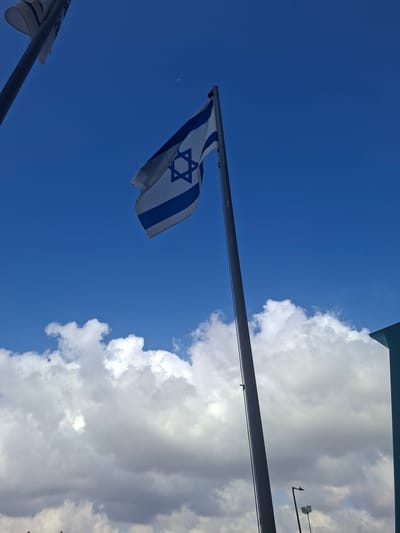 A tall flagpole stands in the foreground, flying the Israeli flag. The flag is predominantly white with two blue horizontal stripes and a blue Star of David in the center. The sky is a clear, bright blue with scattered white clouds. In the background, a part of a light blue building is visible on the right. The scene appears to be outdoors, possibly in an open area within the Shomron Regional Council, Palestinian Territories. There are no people or animals visible in the image.