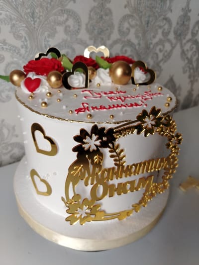 A round, white frosted cake is decorated with gold accents. The top features red roses, green leaves, white meringue swirls, gold spheres, and black heart shapes. The sides are embellished with gold heart cutouts and a gold floral wreath surrounding Cyrillic lettering. The inscription on top is written in red icing. The cake sits on a slightly larger, white-covered base.
