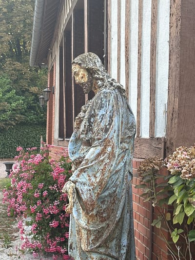 The image captures an aged, weathered statue of Jesus Christ positioned outdoors, to the left of a building with half-timbered architecture. The statue, appearing to be made of metal with a chipped and rusted blue patina, depicts Jesus in a contemplative pose, with his head bowed and his hands clasped. Behind the statue, a vibrant bush of pink flowers, likely geraniums, cascades down. To the right of the statue, the building's brick facade is visible, adorned with vertical wooden beams and white plaster infill. Dry, faded hydrangeas are also present near the statue's base. The background reveals lush green foliage and trees, suggesting a natural, possibly rural or garden setting. The overall impression is one of aged beauty and tranquility.