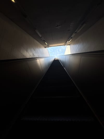 The image depicts an upward-looking view from the bottom of a dark escalator tunnel, located in the city of Bağcılar, Turkey. The dominant feature is a dark, upward-moving escalator, its steps and lower section largely obscured by shadow. The escalator's side panels, also dark, show a subtle glint of reflected light towards the upper right, indicating their reflective surface.
On both sides of the escalator, the tunnel walls are lined with light-colored, possibly beige or off-white, rectangular tiles arranged in a grid pattern, extending upwards. The ceiling, dark and appearing concrete, is equipped with multiple linear fluorescent light fixtures, several of which are illuminated, casting a faint glow within the tunnel.
At the top of the escalator, a brightly lit rectangular opening serves as the exit. Through this opening, a clear blue sky with sparse white clouds is visible, indicating an outdoor, daylight environment. Against the sky, two distant streetlights or utility poles can be observed. The scene is devoid of any individuals, specific identifying signs, or extraneous objects, presenting a minimalist view of an urban transition point.