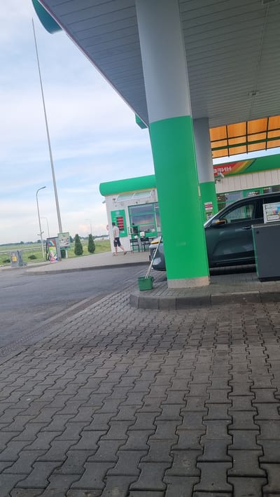 A person wearing a white t-shirt and dark shorts is walking away from a gas station building. The person is walking on a paved area in front of the building. The gas station building is green and white and has a sign with Russian lettering that appears to say "БЕНЗИН," which translates to "GASOLINE." A dark-colored car is parked next to the building. A green bucket with a mop is also visible near the building. The scene is outdoors, and the sky is cloudy. The ground is paved with interlocking bricks. In the background, there are streetlights and trees. The location is identified as Валожынскі сельскі Савет, Belarus.