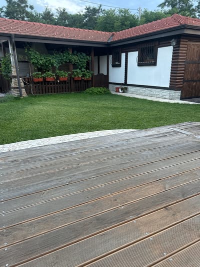 The image captures a view of a residential property with a wooden deck in the foreground, a well-maintained lawn, and a building with a tiled roof in the background. The building appears to be a house or a guesthouse, featuring a mix of white-washed walls and wooden log construction. There are decorative iron bars on the windows, and the property is surrounded by greenery, including climbing plants and trees. The scene suggests a pleasant outdoor living space. The location is identified as Войводино, Bulgaria.