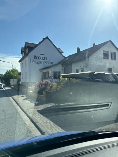 The image shows a building with the text "BRÜCKEL'S HOLZOFENBROT" on its facade. The building appears to be a bakery, as "Holzofen Brot" translates to "wood-fired bread". The building is white with dark lettering and has several windows. There are flowers planted in front of the building. The building is situated on a street with a sidewalk made of paving stones. The sky is blue and clear, suggesting a sunny day. The sun is high in the sky, indicating it is likely midday or early afternoon. The scene is a street in what appears to be a residential or commercial area in Germany, given the text on the building and the architectural style. There are no people visible in the image. The overall mood is calm and ordinary, depicting a typical street scene on a bright day.