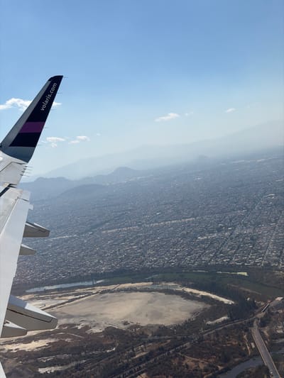 An airplane wing with a "volaris.com" logo is visible in the lower left of the frame, partially obscuring a cityscape and mountain range in the distance. The city is dense with buildings, hazy in the sunlight. Adjacent to the city, a large dry lakebed with sparse vegetation runs alongside what appears to be a road or railway. The sky is blue with some small clouds. The overall scene is an aerial view, likely taken from the airplane itself.