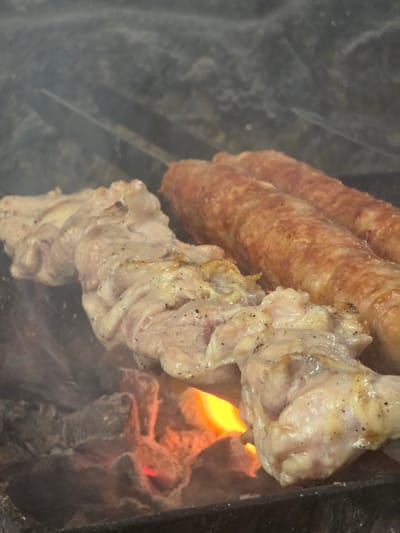 The image shows food cooking over an open flame. The main subjects are several sausages and pieces of chicken, skewered and arranged vertically. The sausages are elongated and have a reddish-brown, slightly textured casing, suggesting they are grilled or roasted. The chicken pieces are lighter in color, with visible seasoning, and appear to be partially cooked, with some parts showing browning.
The scene is set in an outdoor cooking environment, likely a barbecue or grill. Embers and flames are visible beneath the food, indicating the heat source. Smoke is also present, contributing to the atmosphere of cooking. The background is dark and somewhat indistinct, possibly suggesting a cooking pit or a grill enclosure.
The activity taking place is the grilling or roasting of meat. The food is positioned close to the heat source, implying it is in the process of being cooked. There are no people visible in the frame, so the focus is solely on the food and the cooking process.
Notable details include the texture of the sausages and the way the chicken is seasoned. The bright orange and red hues of the flames contrast with the muted tones of the meat and the dark background. The presence of smoke adds a sensory element to the visual. No visible text is present in the image. The context provided is Pidhorodne, Ukraine, which suggests this is likely an outdoor meal preparation in that location.