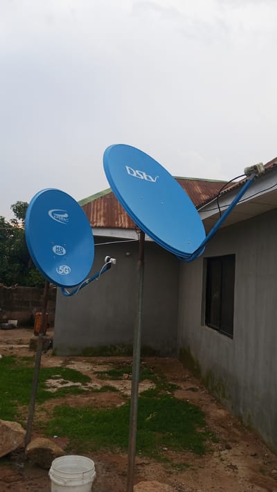 The image depicts an exterior view of a building with two blue DStv satellite dishes. One dish is mounted on a pole in the foreground, and the other is attached to the side of the building via an arm. The building has a corrugated metal roof and a grey concrete exterior wall with a dark-framed window. The ground is a mix of dirt, patches of grass, and stones. A white bucket is visible on the ground in the lower left corner. The sky is overcast. The scene is located in Barikin Sabon Gidan Kanat, Nigeria.