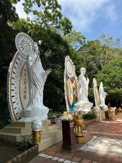 The image displays a serene outdoor setting with several large white statues of Buddhist figures. The statues are ornate, with one prominently featuring a large, decorative halo and holding a lotus flower. The statues are positioned along a paved walkway made of red bricks and patterned tiles, leading into a lush green environment with dense trees and foliage. The sky above is blue with scattered white clouds. Several golden urns and offering boxes are placed near the statues. The overall scene suggests a temple or a spiritual site located in a natural, verdant area. While the specific location is in Thuận Nam, Vietnam, the image itself doesn't provide visual cues to pinpoint a precise landmark within the city.