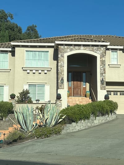 The image shows a large, two-story house with a beige stucco exterior and a tile roof. The house features a prominent arched entryway with stone accents, leading to a double wooden front door. To the left of the entrance, there are several windows with light blue shutters. To the right, there is a garage with a beige door. The front yard is landscaped with a variety of plants, including large agave plants, green shrubs, and flowering bushes. A gray driveway leads up to the garage. The sky is clear and blue. The house is situated on a slight incline. The image appears to be taken on a sunny day. The location is identified as Rocklin, United States of America. There are no people or animals visible in the image.