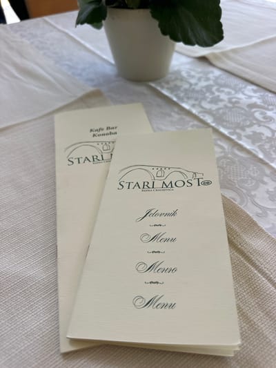 The image shows two menus on a table set with a white, textured tablecloth. One menu is partially obscured by the other. Both menus are a light cream color and feature elegant, dark script. The top menu displays a logo with a bridge and the text "STARI MOST@" and "RIJEKA CRNOJEVIĆA". Below the logo, the word "Jelovnik" is prominently featured, followed by "Menu" in a slightly smaller font. The second menu, peeking out from underneath, also has a similar logo with "STARI MOST" and "RIJEKA CRNOJEVICA" and the text "Kafe Bar Konoba". The menus are angled on the table, and a green leaf from a plant is visible in the lower left corner, suggesting a restaurant or cafe setting. The lighting appears to be natural, indicating it might be daytime. The overall impression is one of a well-presented dining establishment in Rijeka Crnojevića.