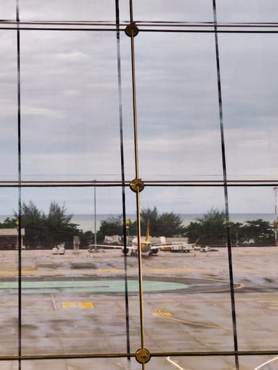 This image, taken from inside an airport terminal through a large glass window, shows an airport tarmac with a passenger airplane parked at a gate. The airplane has a white fuselage with a yellow and red tail. Several ground service vehicles, including a baggage cart and a service truck, are positioned around the aircraft. The tarmac is wet, indicating recent rain, with yellow and green markings visible on the surface. In the background, a line of trees separates the tarmac from the ocean, which appears calm under an overcast sky. The view is partially obstructed by vertical and horizontal metal mullions and fixtures that are part of the window structure. The scene is located in Tambon Mai Khao, Thailand, likely at Phuket International Airport, given its proximity to the sea and tropical vegetation. No people are clearly visible in the image.