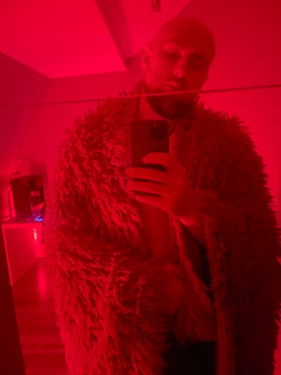A person is taking a selfie in a dimly lit room with red lighting. The person is bald with a beard and is wearing a fluffy, shaggy jacket or coat. They are holding a smartphone in their right hand, partially obscuring their face. In the background, a mirror reflects the scene, and to the left, there's a glimpse of a shelf with a black object, possibly an electronic device, illuminated by a blue light. The environment appears to be indoors, with walls and a ceiling visible. No specific location in Gliwice, Poland can be identified from the image.