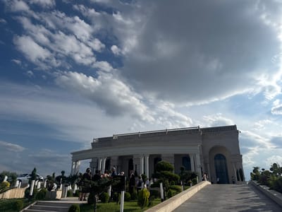 This is a media file depicting an outdoor scene in Mkhchyan, Armenia, likely during the daytime. The sky is filled with a dramatic display of clouds, with patches of blue sky visible, suggesting a day with mixed weather conditions. Sunlight breaks through, creating varied lighting across the scene.

The main subject is a grand, neoclassical-style building with a portico supported by columns, a balustrade along the roofline, and large arched windows. In front of the building, a group of people are gathered, some dressed in formal attire, suggesting a special occasion or event. The landscaping includes manicured bushes, trees, and a paved pathway leading up to the building. A white luxury car is parked in the background on a raised level surrounded by greenery and fountains.

The overall atmosphere appears to be one of a gathering or celebration. The building's architecture and the presence of guests suggest it might be a venue for events like weddings or official functions. The lighting and cloud formations create a somewhat dramatic and possibly awe-inspiring visual effect. No visible text is present in the image.