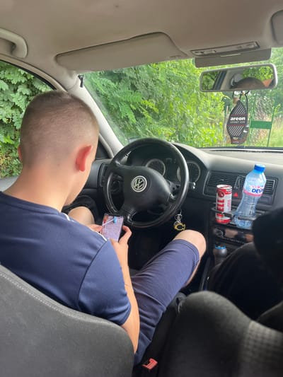 In the driver's seat of a car, a young person is holding a smartphone horizontally. The person is wearing a navy blue t-shirt with white stripes on the sleeves and has short, light-brown hair. Their right arm is resting on their leg, and their left arm is bent as they hold the phone. The car's interior is visible, including the steering wheel with a Volkswagen logo, the dashboard with an air vent, a water bottle, and a can of "100 RED GRAPE" drink. A keychain with a silver engine-shaped charm hangs from the steering column.
The scene appears to be set during the daytime, with natural light filtering into the car. The background outside the windows shows lush green foliage, suggesting a rural or natural setting. A rear-view mirror is visible on the left, reflecting a portion of the car's interior and some greenery. A "areon SPORT LUX SILVER" air freshener is hanging from the mirror. The overall mood seems relaxed, with the person engaged with their phone. The location is Madzherito, Bulgaria.