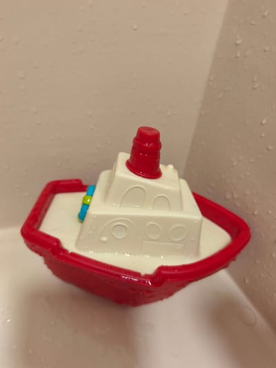 A red and white toy tugboat sits in a shallow pool of water, likely in a bathtub. The tugboat has a red smokestack and a blue and yellow detail on its deck. The walls of the bathtub are a light beige color and are speckled with water droplets. The scene is brightly lit. This image was taken in Platja D'aro, Spain.