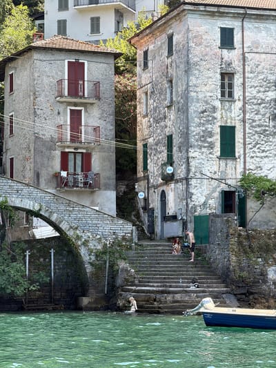 The image depicts a scene in Nesso, Italy, featuring old, weathered buildings situated along a body of water. On the left, a multi-story building with red shutters and balconies stands adjacent to a stone bridge arching over the water. Steps lead up from the water's edge to the buildings. In the foreground, the turquoise water is visible, with a small blue boat docked to the right. Several people are present: one person is partially submerged in the water near the steps on the left, and a group of three individuals are sitting and standing on the steps further up. The buildings themselves show signs of age, with peeling paint and textured facades. Lush green foliage surrounds the structures, indicating a natural, somewhat overgrown environment. The overall impression is of a historic lakeside village.