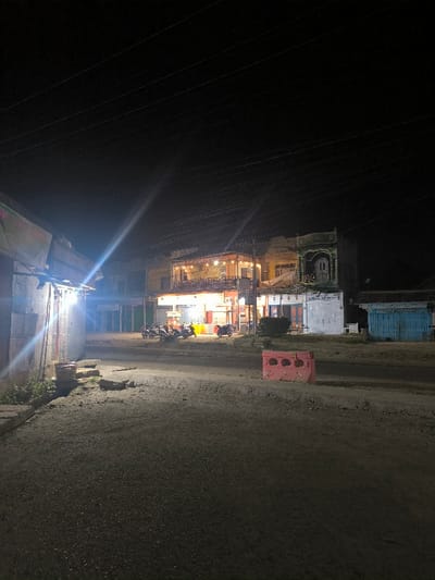 The image depicts a nighttime street scene in Manetunong, Indonesia. The foreground consists of an unpaved or poorly maintained road surface. A red plastic barrier is positioned centrally on the right side of the road. To the left, a single-story structure is brightly illuminated by an unseen light source, casting a significant lens flare; an overhead tarp is visible. In the midground, a two-story building complex, brightly lit with warm and white artificial lights, stands on the right side of the road. This complex appears to house commercial establishments, with a balcony on the upper floor. Multiple motorcycles are parked in front of this building. Adjacent to it on the right is another two-story building with a distinct green-colored upper facade and closed lower-level shutters. Further right, a small, light blue single-story structure is present. The background is a dark night sky, traversed by multiple overhead power lines. No people are visible within the scene.