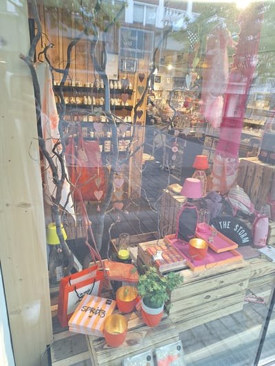 The image shows a shop window display, likely in Bochum, Germany, given the context provided. The display features a rustic theme with natural elements and a variety of decorative items. In the foreground, a wooden crate forms a base for several objects. A bright orange shopping bag with black handles sits on the left, next to a stack of striped boxes with the word "SPRITZ" printed on them. Two small, orange, candle-holder-like objects with gold interiors are placed below the boxes, one of which contains a lit candle. A potted plant with small white flowers is to the right of the "SPRITZ" boxes.

Above these items, a pink tray holds a pink candle and a small glass container. To the right of the pink tray, a black fan-like object with white lettering that reads "THE STORM" is visible. Further to the right, a pink bottle and a pink bag with white lettering are displayed. In the background, the shop interior is visible, with shelves filled with small bottles or containers. A large, dark, bare branch structure dominates the center of the display, with decorative heart shapes and other small ornaments hanging from it. Two lamps with pink and orange shades are also present in the mid-ground. Reflections from the outside world are visible on the shop window, including trees and a building.