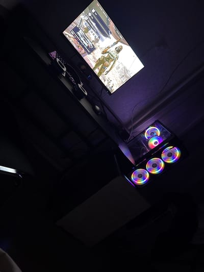 This is a dimly lit, angled photograph of a gaming setup. The main focus is a computer tower with multiple RGB fans, emitting a rainbow of colors. Above the tower, a monitor displays a video game with a character in tactical gear. A pair of headphones rests on a surface in front of the monitor. The scene appears to be indoors, possibly in a room where the primary light source is the computer's illumination. There are no visible people or interactions. The environment is cluttered with cables and components of the gaming setup. The image has a purplish hue, likely from ambient lighting.