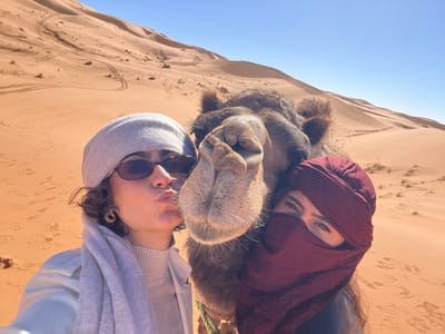This close-up selfie captures two individuals and a brown Dromedary camel positioned against a backdrop of extensive sand dunes under a clear blue sky in the desert near Rissani, Morocco. On the left, a female with short, curly brown hair wears dark sunglasses, a light grey beanie, a light-colored turtleneck, a light beige jacket, and a light grey scarf. She is making a puckered "kissing" face, directing it towards the camel's snout. A gold-colored hoop earring is visible on her left ear. The camel's head and neck occupy the central frame, its brown fur visible, and its snout slightly open. To the camel's right, a second female wears a dark reddish-brown traditional headscarf that covers the lower portion of her face, exposing only her eyes. The background consists of towering orange-brown sand dunes exhibiting ripple patterns and scattered tracks.