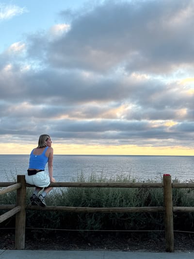 A woman is sitting on a wooden fence overlooking the ocean in Encinitas, California. She is wearing a blue tank top, white shorts, and athletic shoes. She has earbuds in and is looking out at the water. The sky is cloudy with some hints of sunset colors. The fence is made of rough-hewn logs and is covered in greenery. The ocean is calm and stretches to the horizon.