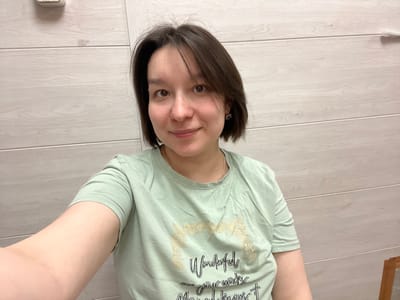 An adult female, appearing to be of East Asian or mixed ethnicity, is depicted taking a selfie indoors. She possesses short, dark brown hair and a light complexion, exhibiting a slight smile while looking directly at the camera. She is wearing a light green short-sleeved t-shirt featuring gold-colored lettering on the chest that includes the word "Wonderful" and additional script. Small, metallic earrings are visible in her ears, and a thin, gold-colored necklace is present around her neck. Her left arm is extended into the lower left foreground, indicative of holding the camera for the selfie. The background consists of a wall clad in light-colored, horizontally aligned panels with a wood grain pattern. In the upper left corner of the background, a small, light-colored rectangular device, possibly a thermostat or sensor, is mounted on the wall. A fragment of a dark, possibly wooden, rectangular object is visible in the bottom right corner, suggesting furniture. The scene is located within a building in the city of Kozlovka, Russia.
