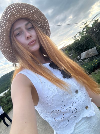 In a selfie taken outdoors, a woman with long, red hair and wearing a straw hat and a white eyelet top poses. Sunglasses are perched on her hair. The background features a cloudy sky, lush green trees, and a glimpse of a hillside. A person in a white shirt and black pants is visible in the distance on a path. The location is identified as Radanovići, Montenegro.