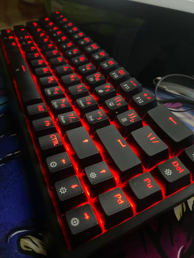 The image shows a close-up, high-angle shot of a black mechanical keyboard with red backlighting. The keys display Cyrillic characters and some English labels such as "FN," "CTRL," "ALT," "F10," "F11," and "F12." The keyboard is resting on a colorful mat or mousepad with a design that is partially visible, featuring blue and purple hues. A pair of eyeglasses with thin metal frames is positioned on the right side of the image, slightly out of focus. The background is dark and appears to be a desk surface, with a hint of a green object in the upper left corner. There are no people or any indication of the city of Nyurba, Russia in the image.