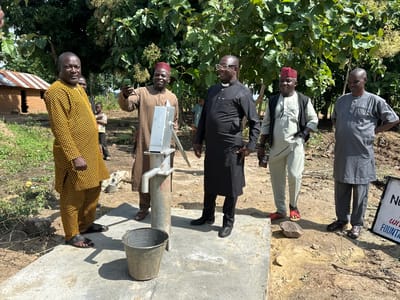 In the city of Afogo, Nigeria, a group of five men stand near a water pump. The man on the left wears a yellow and black patterned tunic and pants. The man next to him, wearing a tan outfit and a red cap, holds up a cell phone, possibly taking a photo or video. To his right, a man in a black cassock with a white clerical collar smiles, and next to him, a man in a light-colored tunic and vest stands with his hands on his hips. The man on the far right wears a grey tunic and pants. A metal bucket sits beside the water pump, which is situated on a concrete platform. In the background, a small mud-brick house with a corrugated metal roof is visible, along with trees and dry, dusty ground.