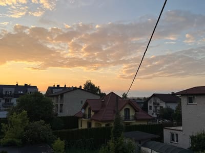 The image captures a sunset over a residential area in Marki, Poland. The sky is filled with soft, golden-orange clouds against a pale blue backdrop, indicating the sun is low on the horizon. Several houses of varying architectural styles are visible. In the foreground, a yellow house with a red tiled roof stands out, featuring balconies and dormer windows. Adjacent to it are dense green hedges and trees. Further in the background, other multi-story buildings with dark roofs and light-colored facades are present, along with more trees and vegetation. A dark diagonal wire runs across the upper right portion of the image. The scene is a quiet, suburban landscape bathed in the warm light of the setting sun. There are no people visible in the image.