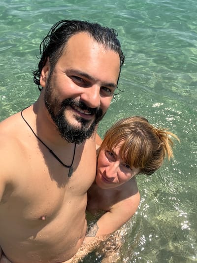 In the city of Sant Pol de Mar, Spain, a couple is captured in a self-taken photograph while submerged in the clear, shallow waters of the Mediterranean Sea. The man, with dark, wet hair and a beard, smiles broadly at the camera. He is wearing a black cord necklace with a rectangular pendant. The woman, with blonde hair tied up, leans her head on his shoulder and looks towards the lens. Her skin is lightly tanned and appears to have some sun redness on her shoulder. Both individuals are wet, with water droplets visible on their skin and hair, indicating they are enjoying a swim. The background consists of shimmering green water, reflecting the bright sunlight.