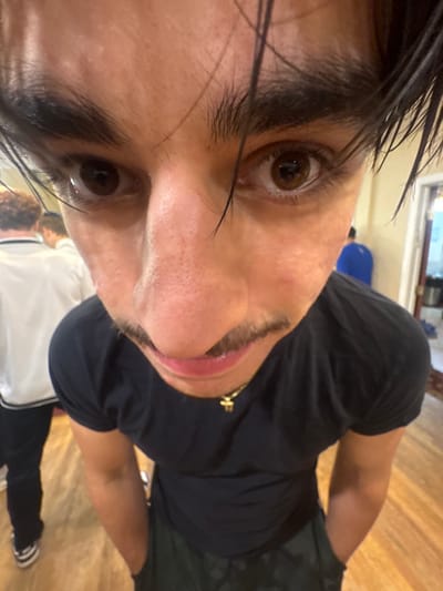 The image displays a close-up, wide-angle perspective of a male individual positioned in the foreground within an indoor setting in Bergenfield, United States. The subject has dark brown eyes, dark eyebrows, and dark hair, with visible strands falling onto his forehead. A thin mustache is present above his upper lip. He is wearing a black short-sleeved t-shirt, dark pants, and a gold-colored necklace with a small cross pendant. His hands are in his pockets, and he is looking directly at the camera. In the blurred background, several other individuals are partially visible: one to the left wears a white long-sleeved shirt with dark trim, and another to the right wears a blue shirt. The floor consists of light-colored wooden planks. An open doorway in the far background reveals indistinct interior elements.