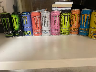 The image shows a collection of Monster Energy drinks lined up on a white surface. The drinks are in various colorful cans, with the iconic Monster Energy claw logo prominently displayed on each. Some of the cans are labeled with names like "Pipeline Punch," "Ultra," "Monarch," and "Mango Loco." The background is slightly blurred, but appears to be an indoor setting with some books stacked in the upper right corner and a curtain in the upper background. The lighting suggests it could be daytime, but the exact time of day is not clear. There are no people visible in the image. The overall scene is one of a display of various flavors of Monster Energy drinks. The visible text on the cans are brand names, flavor names, and nutritional information.