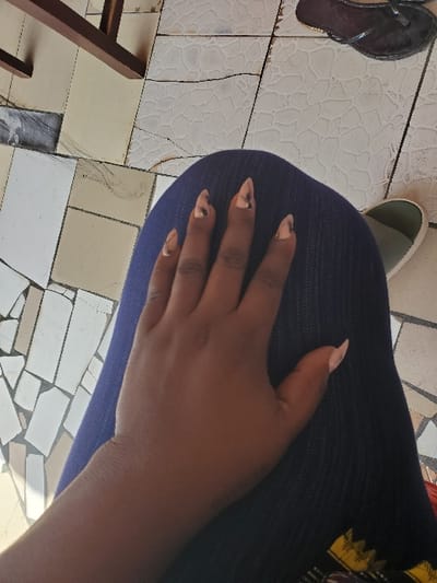 The scene depicts a downward-angled view of a person's left hand resting on their lap, set against a background of tiled flooring. The location is the city of Isunba, Nigeria.

The subject's left hand is dark-skinned, with fingers extended. Four fingers (index, middle, ring, pinky) display elongated, almond-shaped artificial nails featuring a metallic bronze/gold base with irregular dark brown/black patterns. The thumb exhibits a shorter, light pink/beige almond-shaped artificial nail. The hand rests on dark blue fabric, consistent with denim trousers or jeans. A small section of patterned fabric with yellow, black, and red motifs is partially visible on the lower right, below the hand.

The environment consists of an outdoor or semi-outdoor area paved with irregularly shaped and sized ceramic or stone floor tiles in shades of white, beige, and light grey. Many tiles exhibit linear cracking, surface discoloration, and wear. Some light-colored tiles feature a raised, textured pattern. In the upper right background, a partial black thong-style sandal with a dark brown sole is visible. In the lower right background, the front and side of an olive-green and light-colored shoe, possibly a sneaker, are partially visible. In the upper left background, two sections of dark brown wood, likely part of a furniture leg or railing, are present. Natural ambient light illuminates the scene.