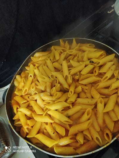 A top-down shot shows a pan filled with cooked penne pasta. The pasta is coated in a yellow or orange-tinged sauce, giving it a uniformly rich color. The pan appears full, with the pasta densely packed. Steam rises faintly from the dish, suggesting it is freshly cooked. The pan sits on a dark surface, possibly a stovetop. Part of the edge of the pan is visible, showing a metallic finish.