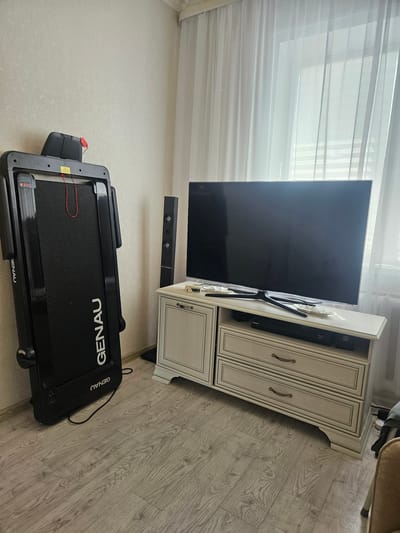The image depicts an indoor scene, likely a living room or bedroom, featuring a black treadmill leaning against a light-colored textured wall on the left side of the frame. The treadmill has the brand name "GENAU" printed vertically in white letters. On top of the treadmill, there is a black control panel with a red safety key attached. To the right of the treadmill, a tall, slender black speaker stands upright. Further to the right, a large flat-screen television is mounted on a white wooden media console with drawers and decorative trim. The television screen is dark, indicating it is either off or displaying dark content. The media console has two drawers visible with antique-style brass handles. A slim black electronic device, possibly a Blu-ray player or game console, is placed on a shelf within the console, partially obscured by the television. A sheer white curtain is drawn across a window to the right of the television, allowing diffused light to enter the room. The floor is covered with light-colored laminate flooring with a wood grain pattern. The overall impression is a domestic interior, tidy and functional, with elements of exercise equipment and home entertainment. The location is not specified in the image.