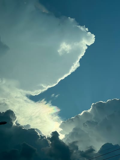 The image captures a dramatic sky over Tambon Don Sak, Thailand, filled with a vibrant display of clouds. The dominant feature is the contrast between bright, sunlit cumulus clouds and darker, shadowed formations. A faint rainbow-like iridescence is visible in the center, suggesting sunlight interacting with water droplets within a cloud. The sky is a deep blue, typical of a clear day, but the cloud formations suggest an impending or recent weather event. In the lower right, thin, dark lines, likely power lines, are faintly visible against the sky, indicating a populated area. The overall impression is one of natural beauty and the dynamic forces of weather.