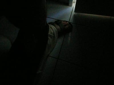 A person is lying on their side on a tiled floor in a dimly lit room. The person is wearing light-colored clothing. A sliver of light from an open doorway illuminates a portion of the scene. The overall impression is one of darkness and low visibility. The image does not contain information that would allow for a specific location in Ümraniye, Turkey to be identified.